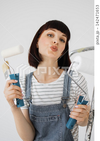 A woman in denim overalls playfully puckers her lips while holding painting tools, ready for a home renovation project A woman in denim overalls playfully puckers her lips while holding painting tools, ready for a home renovation project 142471653