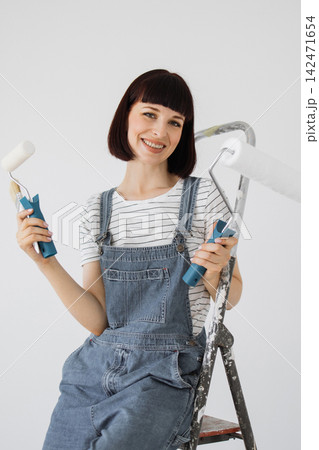 A smiling woman in denim overalls holds paint rollers and a brush while sitting on a ladder, ready for home improvement A smiling woman in denim overalls holds paint rollers and a brush while sitting on a ladder, ready for home improvement 142471654