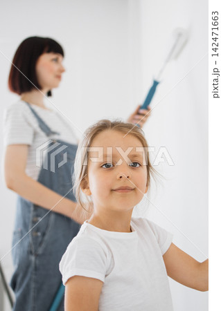 A young girl smiles at the camera while her mother paints the wall in the background with a roller 142471663