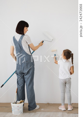 A woman and a young girl stand side-by-side, painting a white wall with rollers, enjoying a home improvement project 142471668