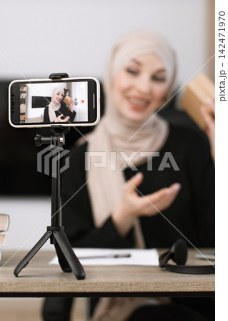 A Muslim woman in a hijab is recording a video blog using her smartphone mounted on a tripod, showcasing a small package 142471970