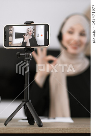 A woman wearing a hijab and headset is recording a video on her smartphone, giving an okay sign 142471977