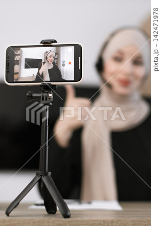 A woman wearing a hijab and headset gives a thumbs-up while recording herself on a smartphone mounted on a tripod, indicating a positive and engaging online presence A woman wearing a hijab and headset gives a thumbs-up while recording herself on a smartphone mounted on a tripod, indicating a positive and engaging online presence 142471978