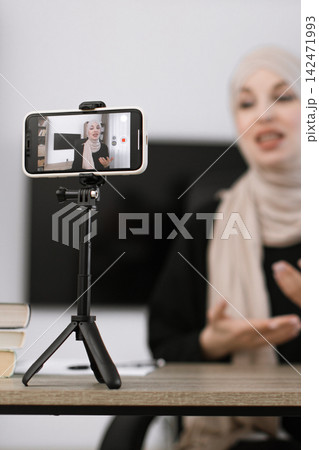 A woman wearing a hijab is recording a video on her smartphone, which is mounted on a tripod, while gesturing with her hands A woman wearing a hijab is recording a video on her smartphone, which is mounted on a tripod, while gesturing with her hands 142471993
