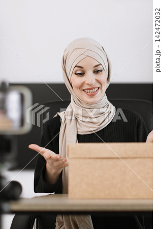 A smiling woman wearing a hijab is unboxing a package while recording a video with her smartphone on a tripod A smiling woman wearing a hijab is unboxing a package while recording a video with her smartphone on a tripod 142472032