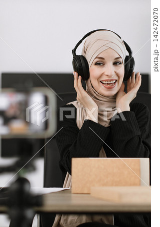 A smiling woman wearing a hijab and headphones is recording content with a smartphone on a tripod, showcasing a modern setup for online creators A smiling woman wearing a hijab and headphones is recording content with a smartphone on a tripod, showcasing a modern setup for online creators 142472070