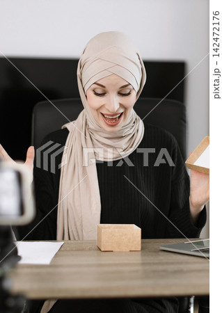 A woman wearing a hijab expresses joy and surprise while opening a small cardboard box in front of a camera, suggesting a live stream or unboxing video A woman wearing a hijab expresses joy and surprise while opening a small cardboard box in front of a camera, suggesting a live stream or unboxing video 142472176