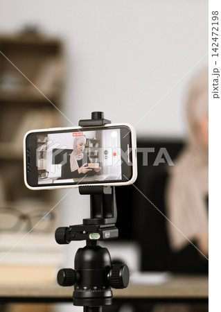 A woman wearing a hijab records herself on a smartphone mounted on a tripod, showcasing a product A woman wearing a hijab records herself on a smartphone mounted on a tripod, showcasing a product 142472198