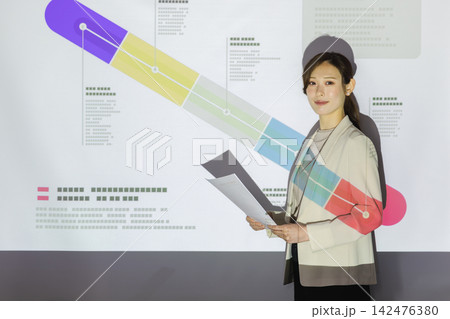 プロジェクターの事業計画画面を背に資料を持ってプレゼンを行う女性ビジネスパーソン 142476380