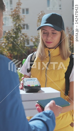 Vertical video. Man open door to a pizza delivery woman wearing yellow thermal backpack with POS wireless terminal for smartphone paying and recieves cardboard boxes of pizza. Food delivery concept. 142478423