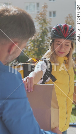 Vertical video. Polite man greets food delivery person with yellow thermal backpack on a bike and receives order or package with food. Food delivery concept. 142478424
