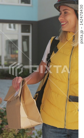 Vertical video. Close up Man customer open door to a food delivery woman wearing yellow thermal backpack on a bike and recieves order or package with food. Food delivery concept. Vertical video. Close up Man customer open door to a food delivery woman wearing yellow thermal backpack on a bike and recieves order or package with food. Food delivery concept. 142478430