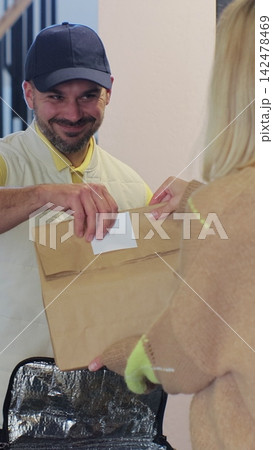 Vertical video. Woman opens door to food delivery man taking order out of thermal insulated bag and giving it to female customer. View from the apartment. Inside shot. Food delivery concept. 142478469