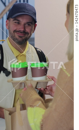 Vertical video. A food delivery person in a yellow uniform. Delivering coffee and food to a person's home. View from the apartment. Inside shot. Food delivery concept. 142478473
