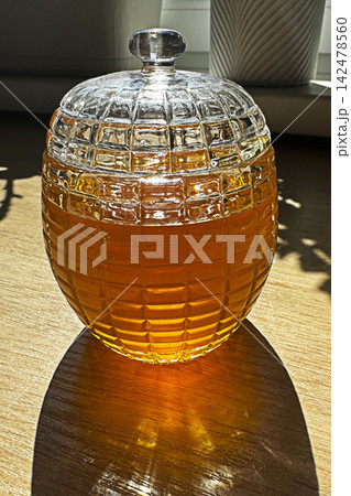 Glass honey jar on wooden table with sunlight casting warm amber shadow in cozy kitchen still life 142478560