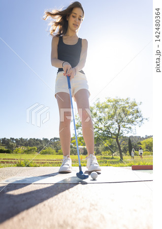 Young woman practices putting at a golf course during bright daytime with clear blue sky and green landscape in the background Young woman practices putting at a golf course during bright daytime with clear blue sky and green landscape in the background 142480364