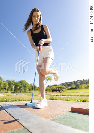 Young girl playing mini golf at a sunny park during the afternoon with blue sky and green landscape in the background 142480390
