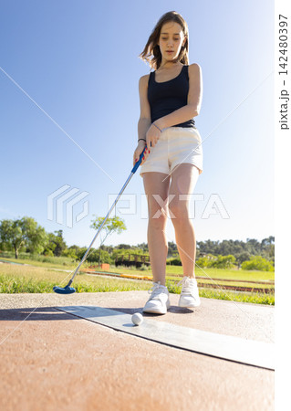 Young woman practices mini golf on a sunny day in a green park with trees in the background Young woman practices mini golf on a sunny day in a green park with trees in the background 142480397