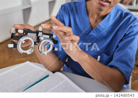 Close-up of medical student hands holding an ophthalmic trial frame over an open textbook. Clinical optometry and refractive diagnostics study Close-up of medical student hands holding an ophthalmic trial frame over an open textbook. Clinical optometry and refractive diagnostics study 142480543