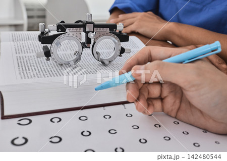 Close-up of a student hand with a pen next to an ophthalmic trial frame and eye test chart. Optometry and visual acuity examination study Close-up of a student hand with a pen next to an ophthalmic trial frame and eye test chart. Optometry and visual acuity examination study 142480544