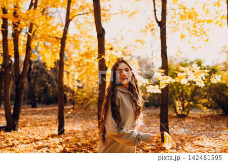Smiling young woman enjoys the autumn weather in the forest with the yellow leaves at sunset. 142481595