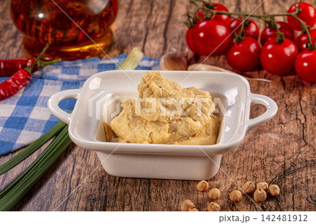 Creamy hummus dip in white bowl, fresh chickpeas, red cherry tomatoes, chili peppers, garlic, green onions, wooden background 142481912