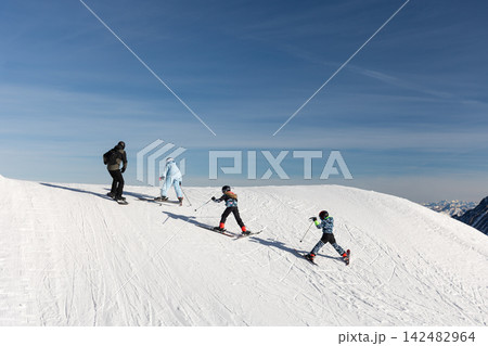 Children skiers climb snowy alpine slope under clear blue sky, showing effort teamwork determination. Challenging ascent symbolizes progress success achievement reaching goals through persistence 142482964