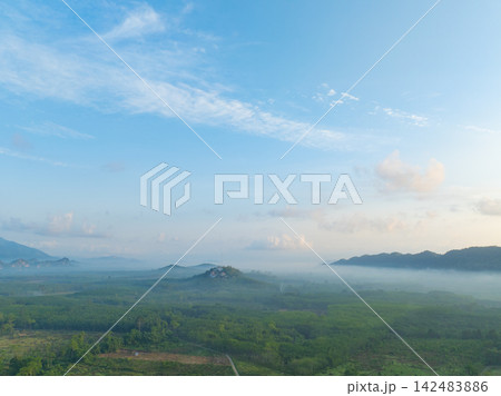 Aerial view of morning mist drifting across tropical valley with forest plantation and distant mountains beneath blue sky. Rural landscape. Tranquil countryside ideal for nature and travel concept. Aerial view of morning mist drifting across tropical valley with forest plantation and distant mountains beneath blue sky. Rural landscape. Tranquil countryside ideal for nature and travel concept. 142483886
