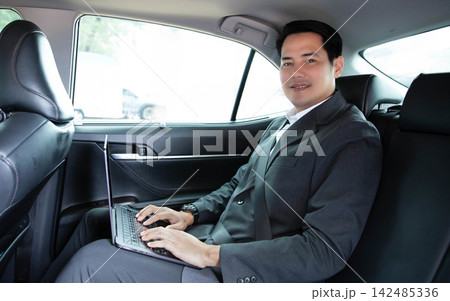 Asian businessman in formal suit smiling while using laptop computer in back seat of car, working remotely during daily commute or business travel, reflecting mobile office lifestyle and success Asian businessman in formal suit smiling while using laptop computer in back seat of car, working remotely during daily commute or business travel, reflecting mobile office lifestyle and success 142485336