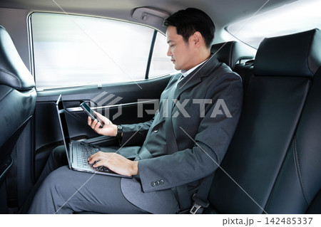 Asian businessman using laptop computer and holding smartphone while sitting in back seat of luxury car, working remotely during daily commute or business travel, reflecting corporate executive Asian businessman using laptop computer and holding smartphone while sitting in back seat of luxury car, working remotely during daily commute or business travel, reflecting corporate executive 142485337