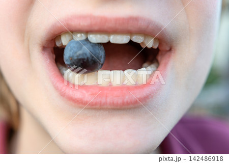 Young person playfully holding a fresh blueberry between their white teeth, representing concepts of childhood nutrition, oral health, and a healthy diet Young person playfully holding a fresh blueberry between their white teeth, representing concepts of childhood nutrition, oral health, and a healthy diet 142486918