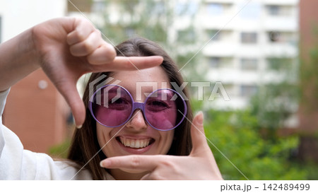 A sweet, cheerful, and charming girl poses for the camera on a city street in the summer. The woman holds up a hand-made frame, like a photograph. A photo frame made by a young hipster girl. A sweet, cheerful, and charming girl poses for the camera on a city street in the summer. The woman holds up a hand-made frame, like a photograph. A photo frame made by a young hipster girl. 142489499