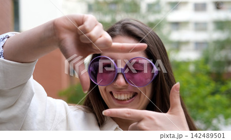 A sweet, cheerful, and charming girl poses for the camera on a city street in the summer. The woman holds up a hand-made frame, like a photograph. A photo frame made by a young hipster girl. A sweet, cheerful, and charming girl poses for the camera on a city street in the summer. The woman holds up a hand-made frame, like a photograph. A photo frame made by a young hipster girl. 142489501