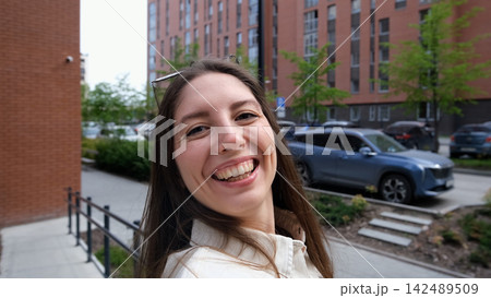 Smiling young woman taking selfie on city steps with modern apartment buildings behind her, casual white shirt, relaxed urban lifestyle, overcast daylight, close portrait view 142489509