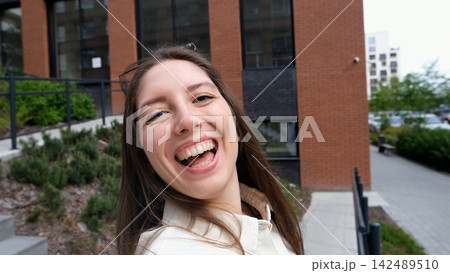 Smiling young woman taking selfie on city steps with modern apartment buildings behind her, casual white shirt, relaxed urban lifestyle, overcast daylight, close portrait view 142489510