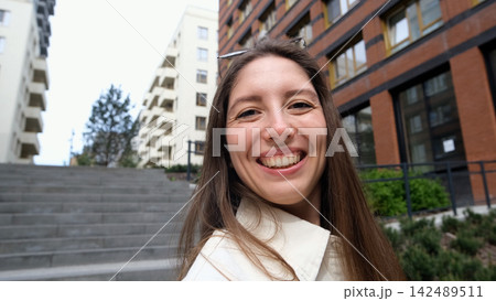 Smiling young woman taking selfie on city steps with modern apartment buildings behind her, casual white shirt, relaxed urban lifestyle, overcast daylight, close portrait view Smiling young woman taking selfie on city steps with modern apartment buildings behind her, casual white shirt, relaxed urban lifestyle, overcast daylight, close portrait view 142489511