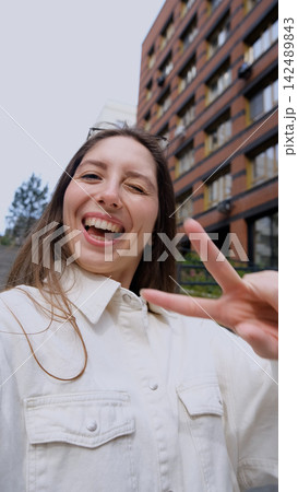 Cute cheerful charming girl poses for camera in summer on city street, hands and fingers showing V sign Cute cheerful charming girl poses for camera in summer on city street, hands and fingers showing V sign 142489843