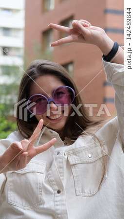 Cute cheerful charming girl poses for camera in summer on city street, hands and fingers showing V sign Cute cheerful charming girl poses for camera in summer on city street, hands and fingers showing V sign 142489844
