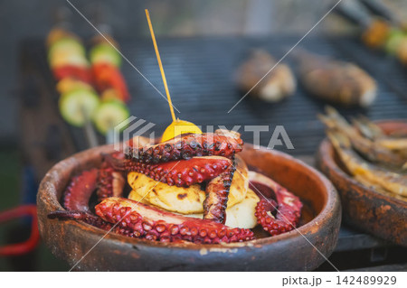 Grilled Octopus Tentacles. Close-up of grilled octopus tentacles with grilled food in rustic ceramic bowl at outdoor food stall. Mediterranean seafood candid scene, culinary and street food themes Grilled Octopus Tentacles. Close-up of grilled octopus tentacles with grilled food in rustic ceramic bowl at outdoor food stall. Mediterranean seafood candid scene, culinary and street food themes 142489929