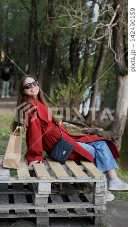 Portrait photo of beautiful young smiling woman in burgundy coat sitting on bench outdoors in park. 142490159