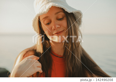 Young woman in a white bucket hat and red top by the sea, looking down with a gentle smile. Warm golden light and a soft coastal background create a natural summer lifestyle portrait with relaxed mood Young woman in a white bucket hat and red top by the sea, looking down with a gentle smile. Warm golden light and a soft coastal background create a natural summer lifestyle portrait with relaxed mood 142490208