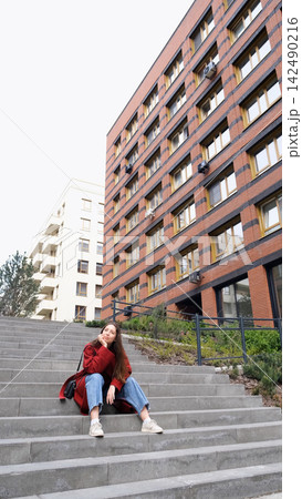 Portrait of a beautiful European woman in a red coat sitting on the steps of a city street 142490216