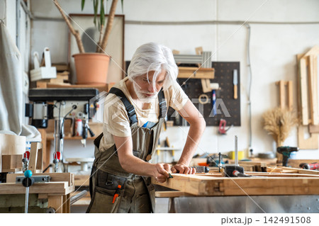 Pensive man restorer sanding surface of wooden door at workbench, restoration workshop workflow 142491508