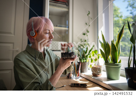 Pink haired woman gently checks zamioculcas cuttings while wearing headphones near window. Pink haired woman gently checks zamioculcas cuttings while wearing headphones near window. 142491532