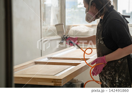 Craftswoman painter wearing protective respirator mask paints antique wooden door with spray gun. 142491860