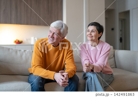 Happy senior married couple husband and wife sitting couch at home looking away at window. 142491863