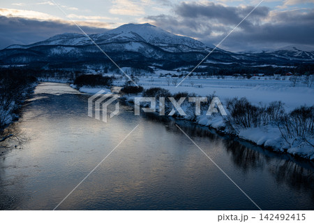 冬の夕暮れ時に輝く尻別川の水面と雪化粧したニセコアンヌプリの雄大な風景 142492415