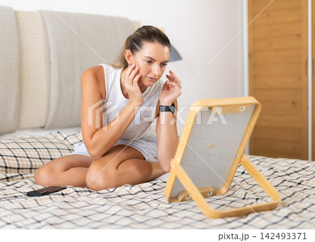 Woman in nightgown looks in the mirror and preens herself in interior of on bed in bedroom Woman in nightgown looks in the mirror and preens herself in interior of on bed in bedroom 142493371