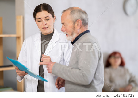 Young nurse or doctor talking with elderly man in reception area Young nurse or doctor talking with elderly man in reception area 142493728