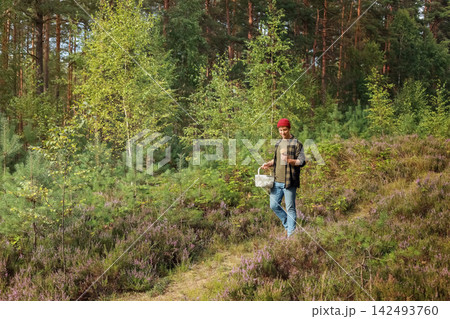 Man walking on a forest path with a basket, picking a wild mushroom. This autumn outdoor activity highlights foraging, seasonal harvest and nature connection. 142493760